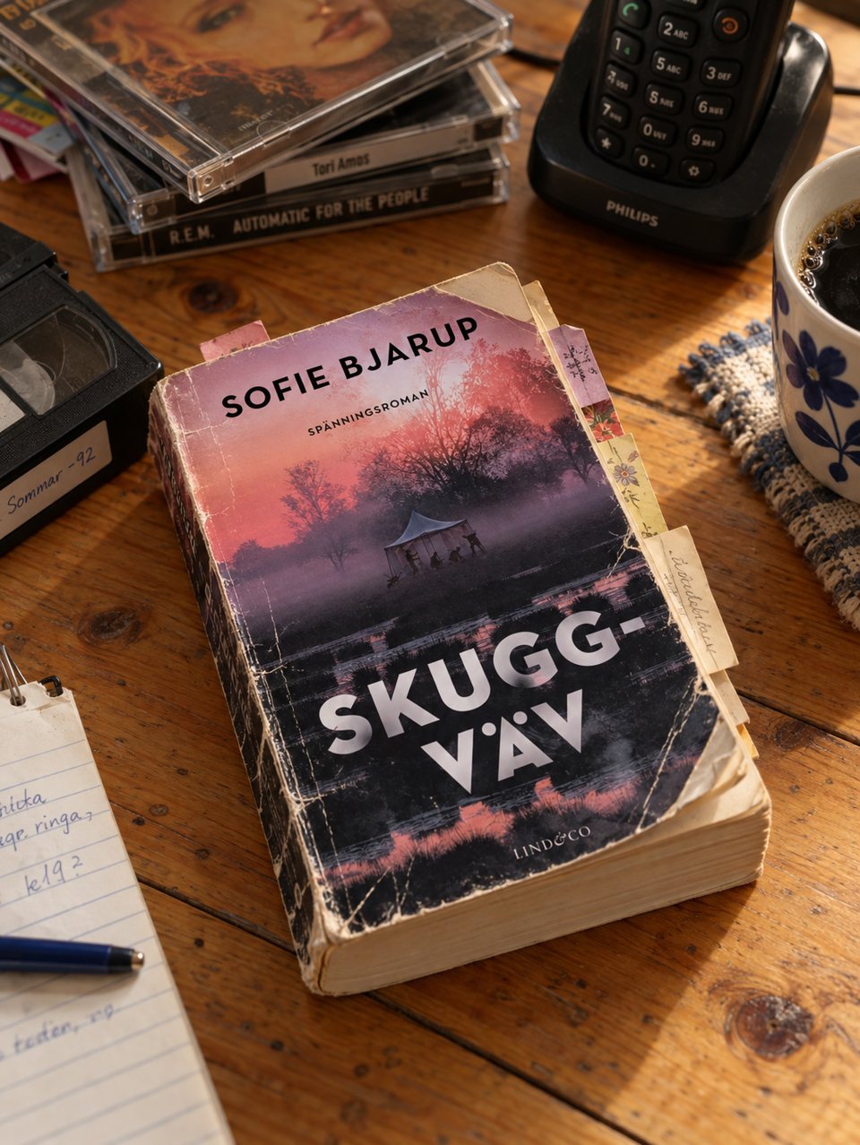 Skuggväv som ett slitet exemplar på ett träbord, med cd-skivor, en videoband märkt Sommar -92, en trådlös telefon, en kaffekopp och ett anteckningsblock