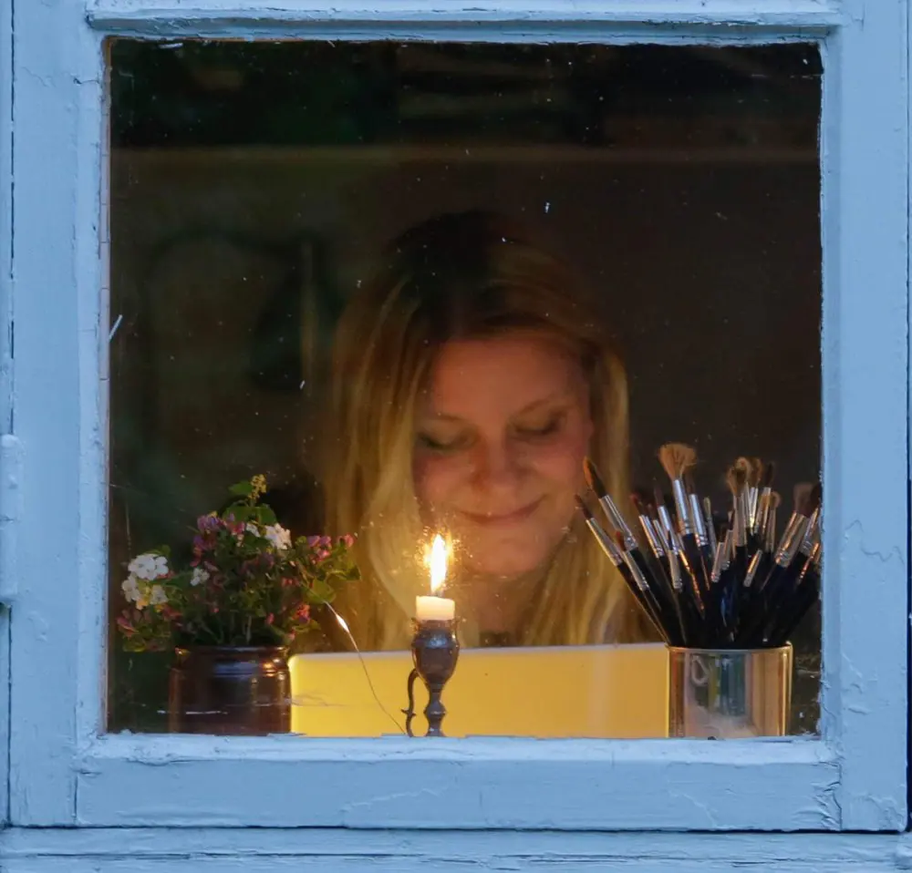 Sofie Bjarup bakom ett fönster, med ett tänt ljus, en blomvas och en burk penslar